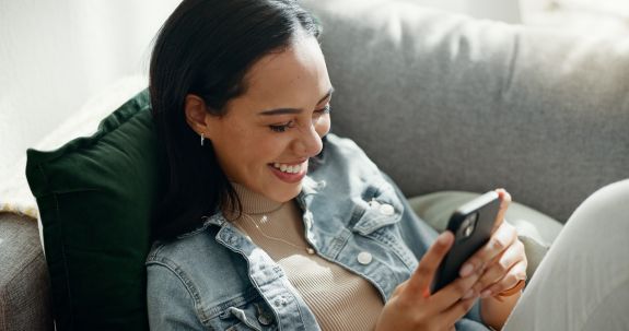 Junge Frau liegt auf dem Sofa und ließt lachend etwas auf dem Handy