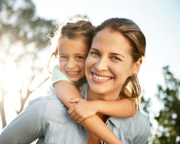 Junge Mutter lacht während sie mit ihrer Tochter huckepack im Freien spielt.